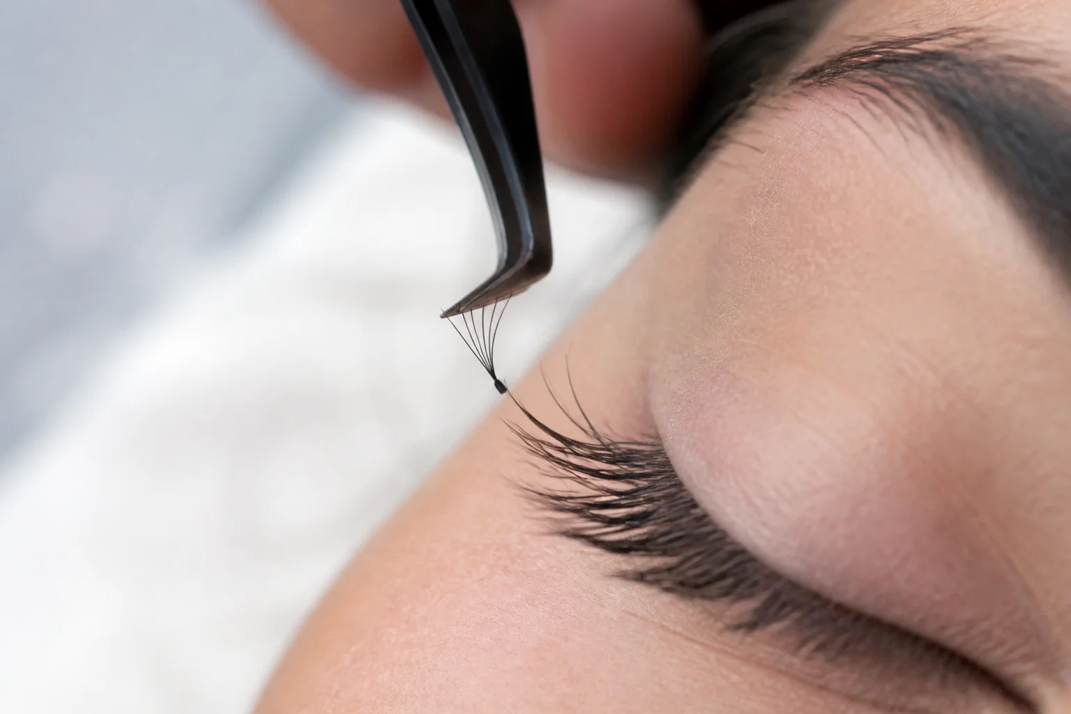 Tweezers placing a small premade fan onto an isolated natural lash beside placed single extensions
