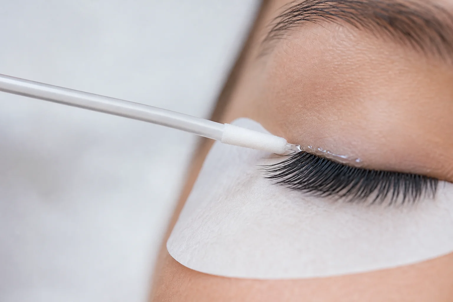 Micro swab applying remover gel along the base of lash extensions on a closed eye with an under-eye pad